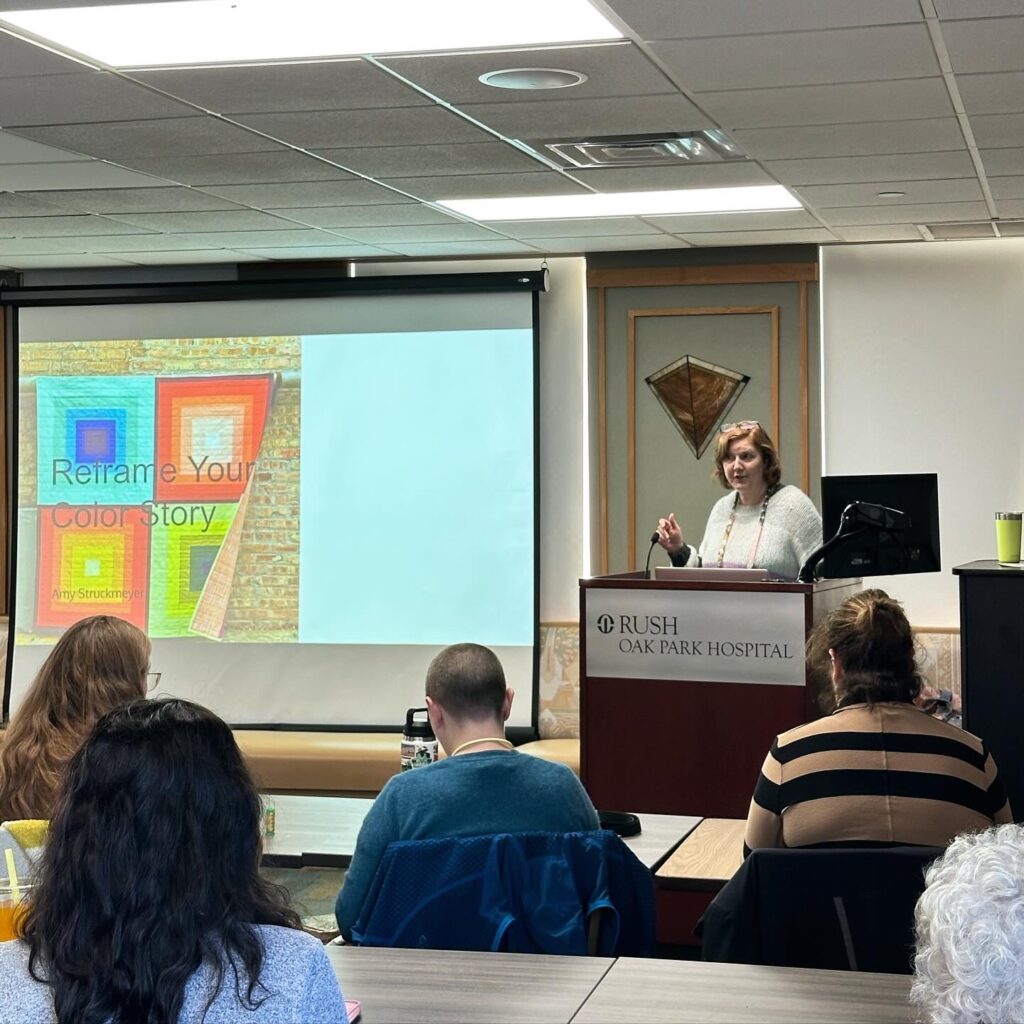 amy, a white woman wearing a white sweater, stands at the front of a crowded room, gesturing toward a colorful powerpoint slide