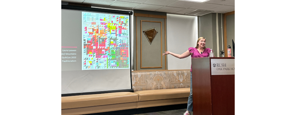 laura loewen, a white woman wearing a pink shirt, gestures at a screen showing one of her quilts